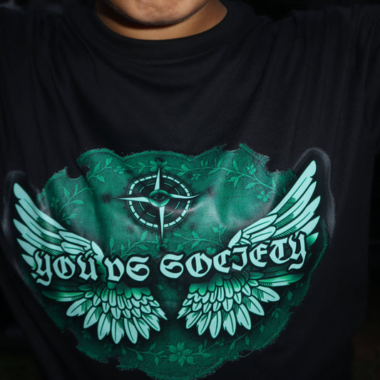 A close-up of a person wearing a black t-shirt with a green graphic print that reads 'You vs Society' with angel wings and a compass target design. 
